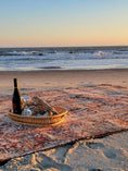 Load image into Gallery viewer, The Ravencourt Rug spread across soft sand on a moody beach, contrasting its vintage pattern with driftwood and surf.