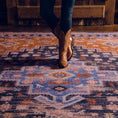 Load image into Gallery viewer, Lifestyle photo of a woman in boots standing on the Twilight Caravan rug, showcasing the vibrant indigo, rust, and violet medallion design
