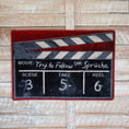 Load image into Gallery viewer, The Cue, Action Rug displayed against a rustic wood floor, highlighting its movie-themed clapperboard design
