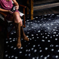 Load image into Gallery viewer, Lifestyle scene with a woman seated cross-legged in a chair above the Nocturna’s Hide rug, showcasing the mysterious pattern beneath soft, ambient light
