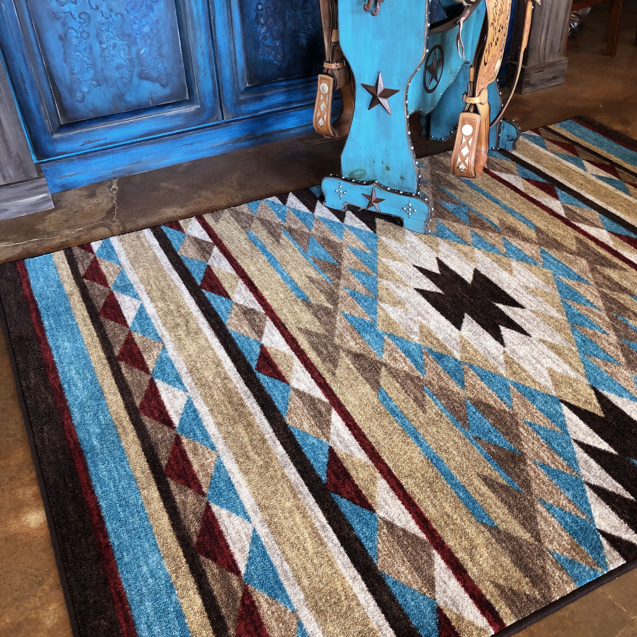 Room scene with the Trail of Glass rug beneath a rustic bench holding a saddle, showcasing the tribal diamond pattern in warm turquoise, gold, and rust tones