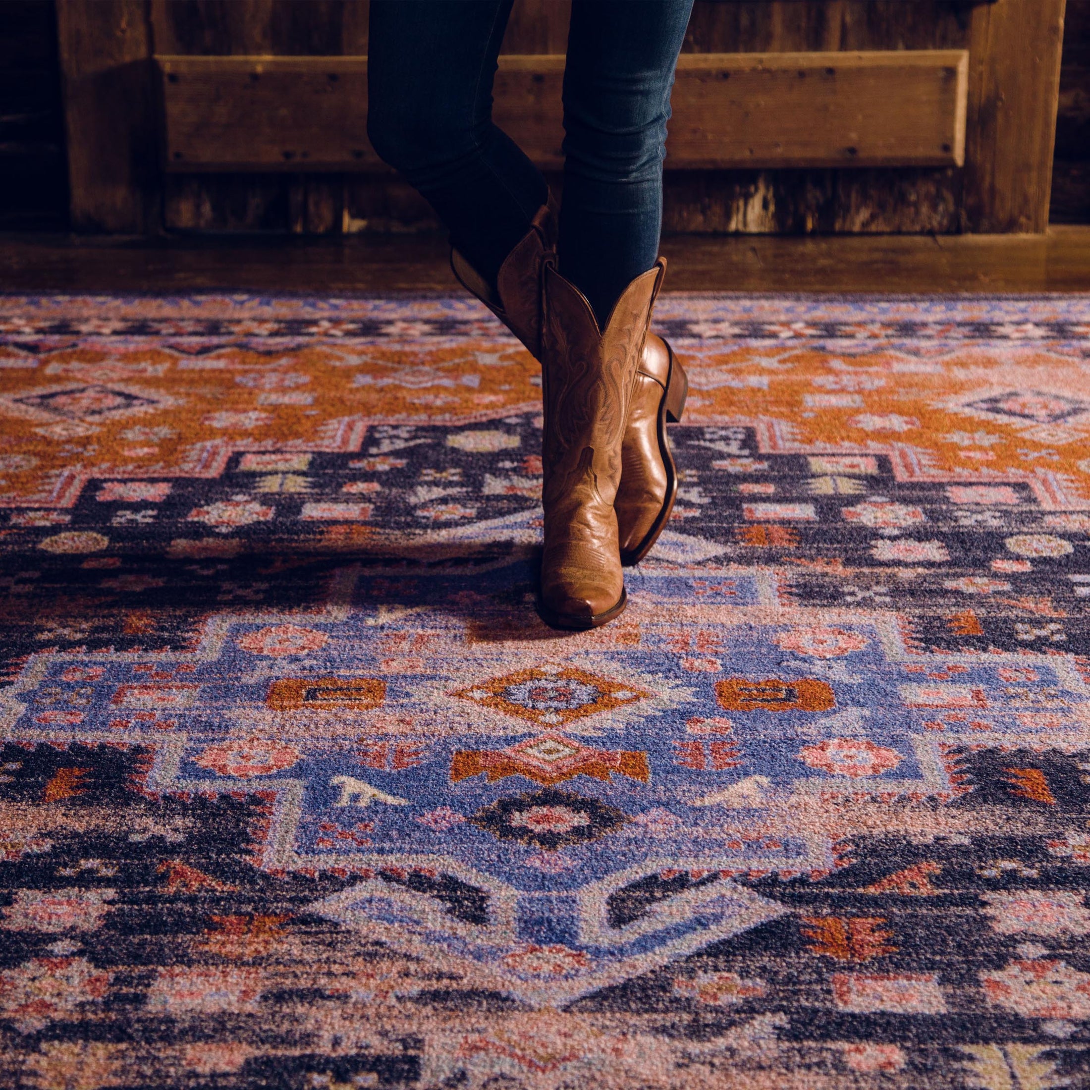 Lifestyle photo of a woman in boots standing on the Twilight Caravan rug, showcasing the vibrant indigo, rust, and violet medallion design

