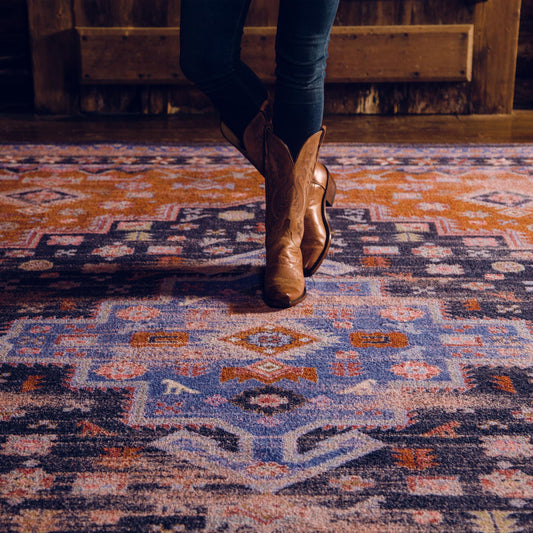 Lifestyle photo of a woman in boots standing on the Twilight Caravan rug, showcasing the vibrant indigo, rust, and violet medallion design

